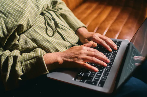 A middle-aged person in a green checkered long-sleeve shirt typing on a laptop indoors, illustrating...