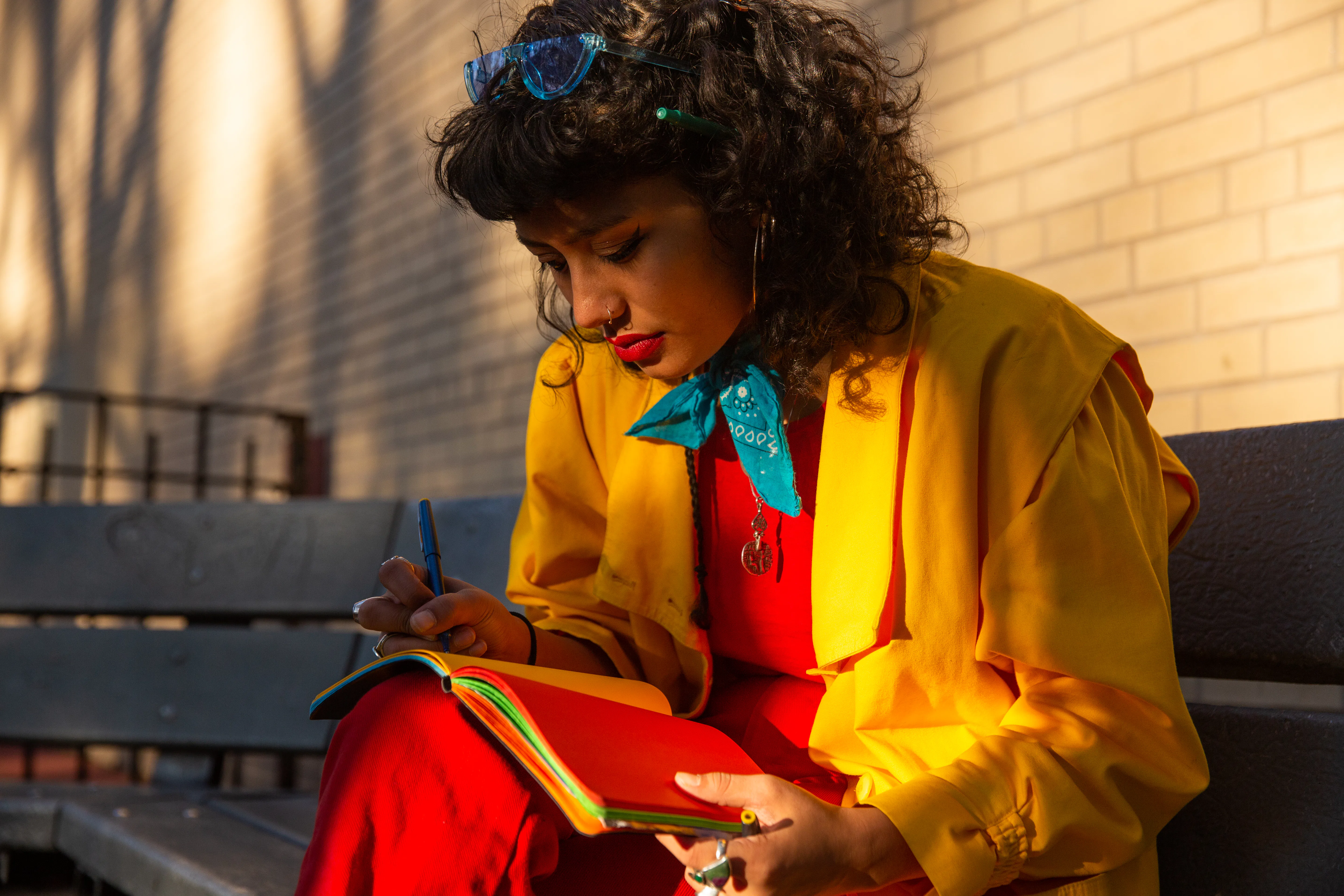 A woman in colorful clothing writes in a journal on a bench. 