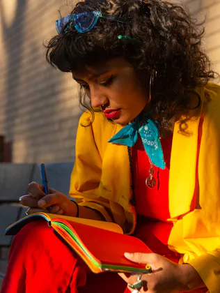 A woman in colorful clothing writes in a journal on a bench.