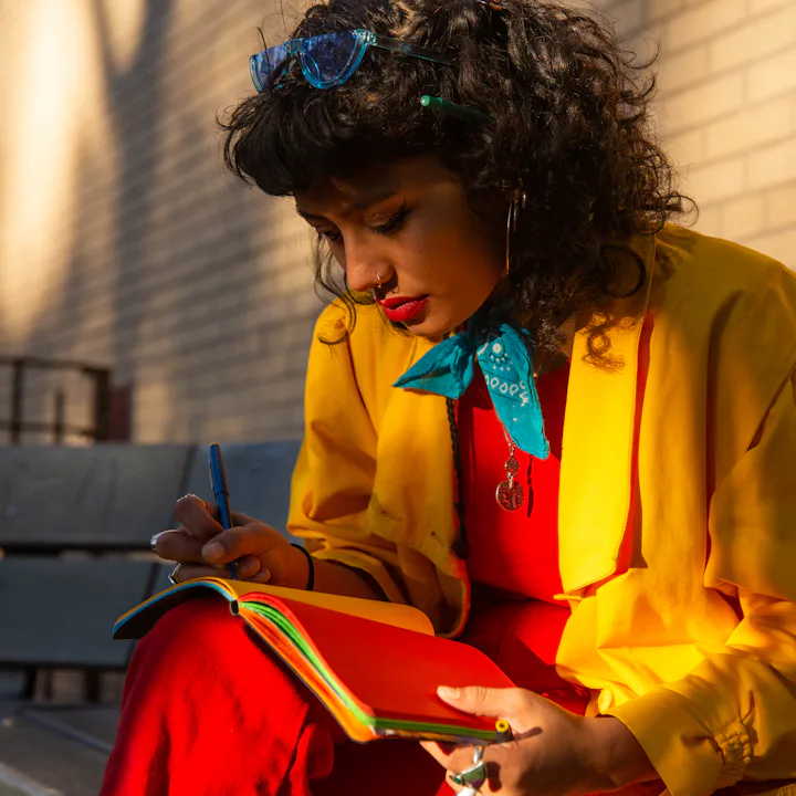 A woman in colorful clothing writes in a journal on a bench.