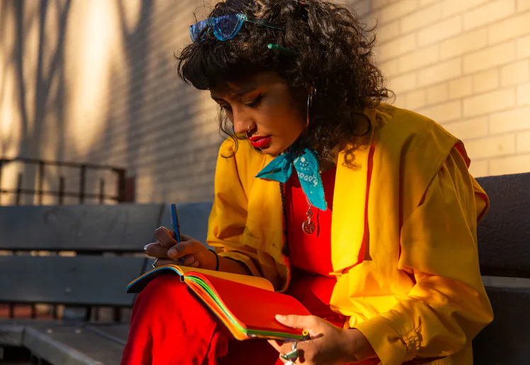 A woman in colorful clothing writes in a journal on a bench.