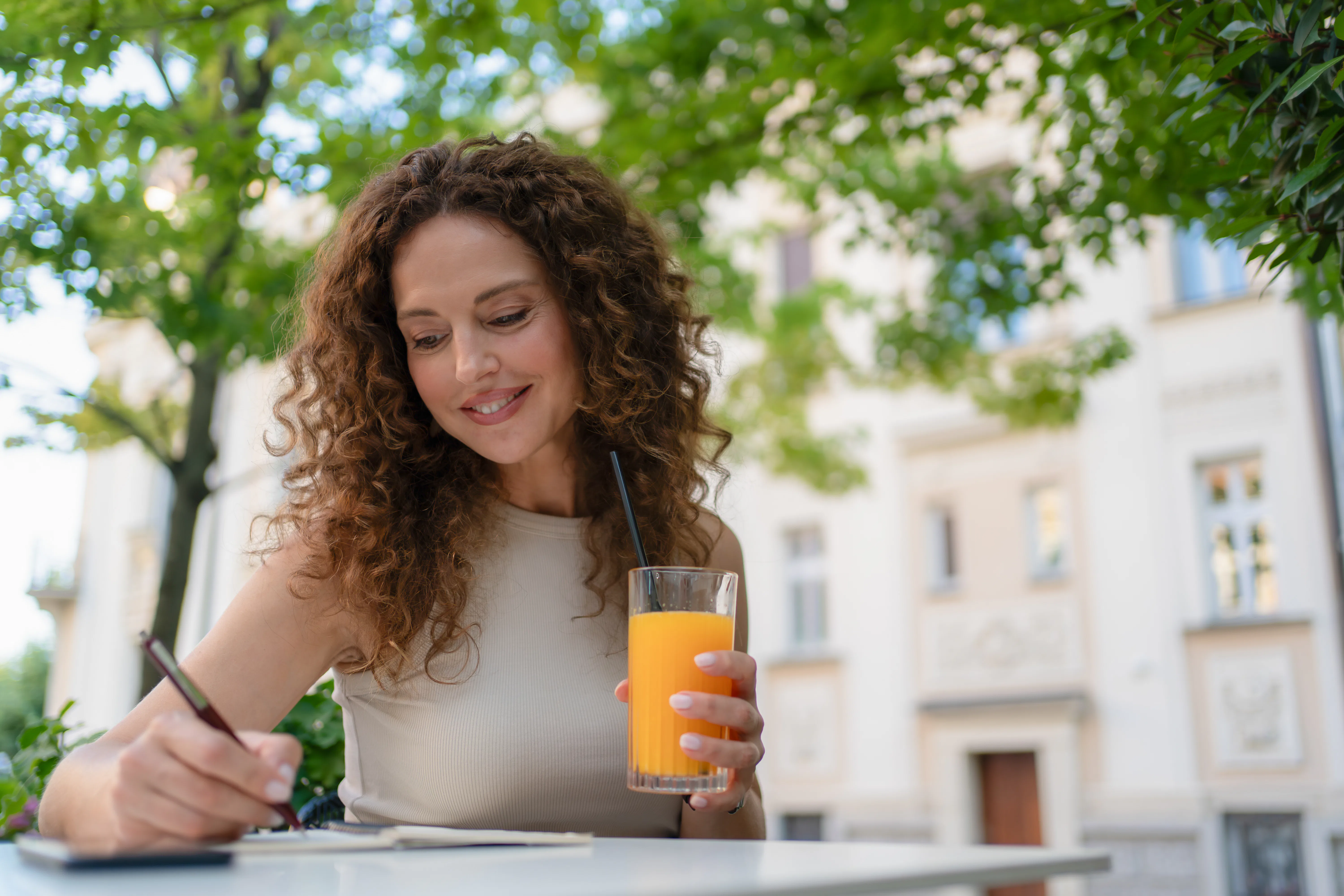Young adult woman writing in notebook enjoying orange juice outdoors
