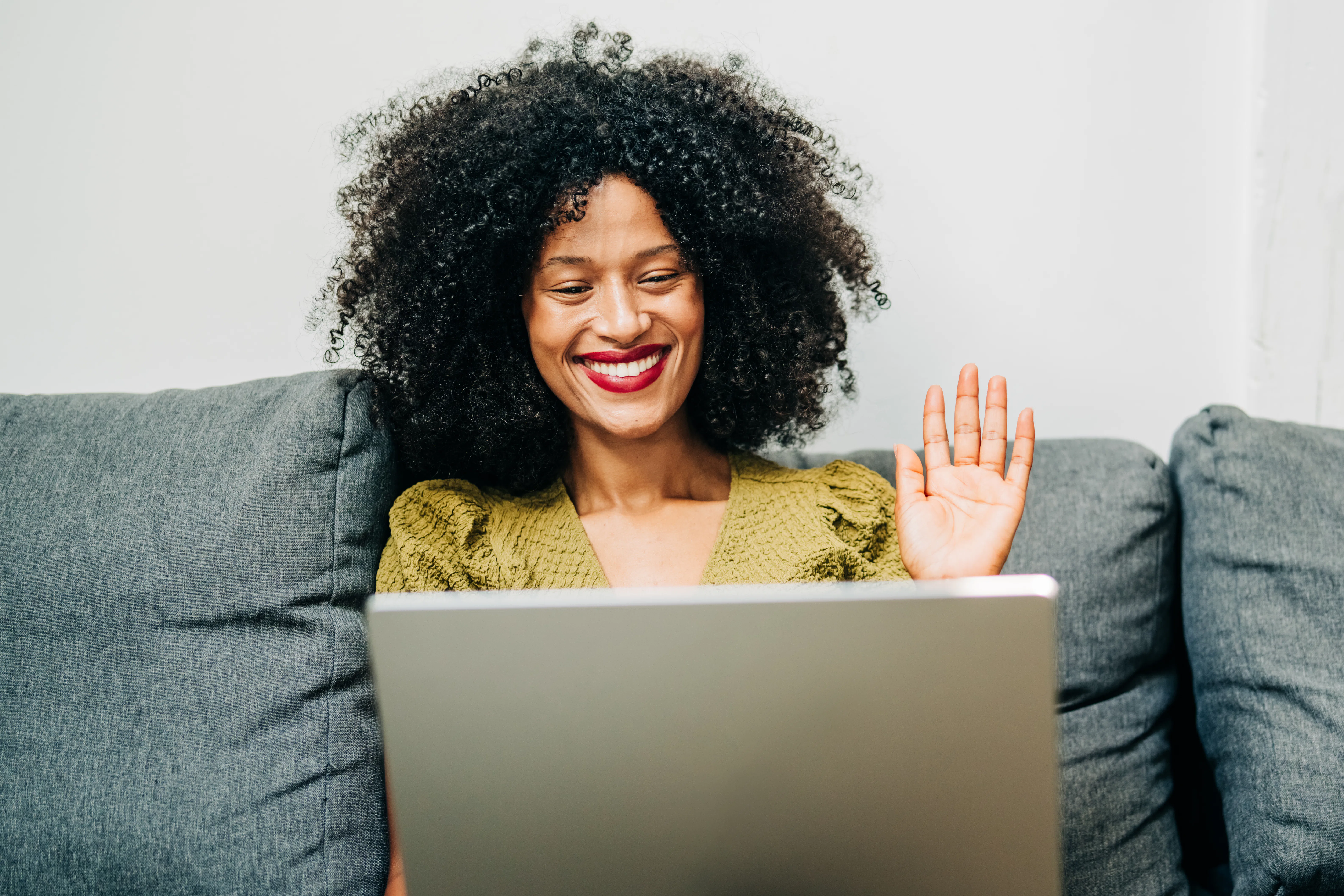 A smiling Black woman with an afro hairstyle waving at her laptop while sitting on a grey sofa. The ...