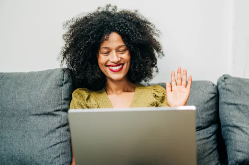 A smiling Black woman with an afro hairstyle waving at her laptop while sitting on a grey sofa. The ...