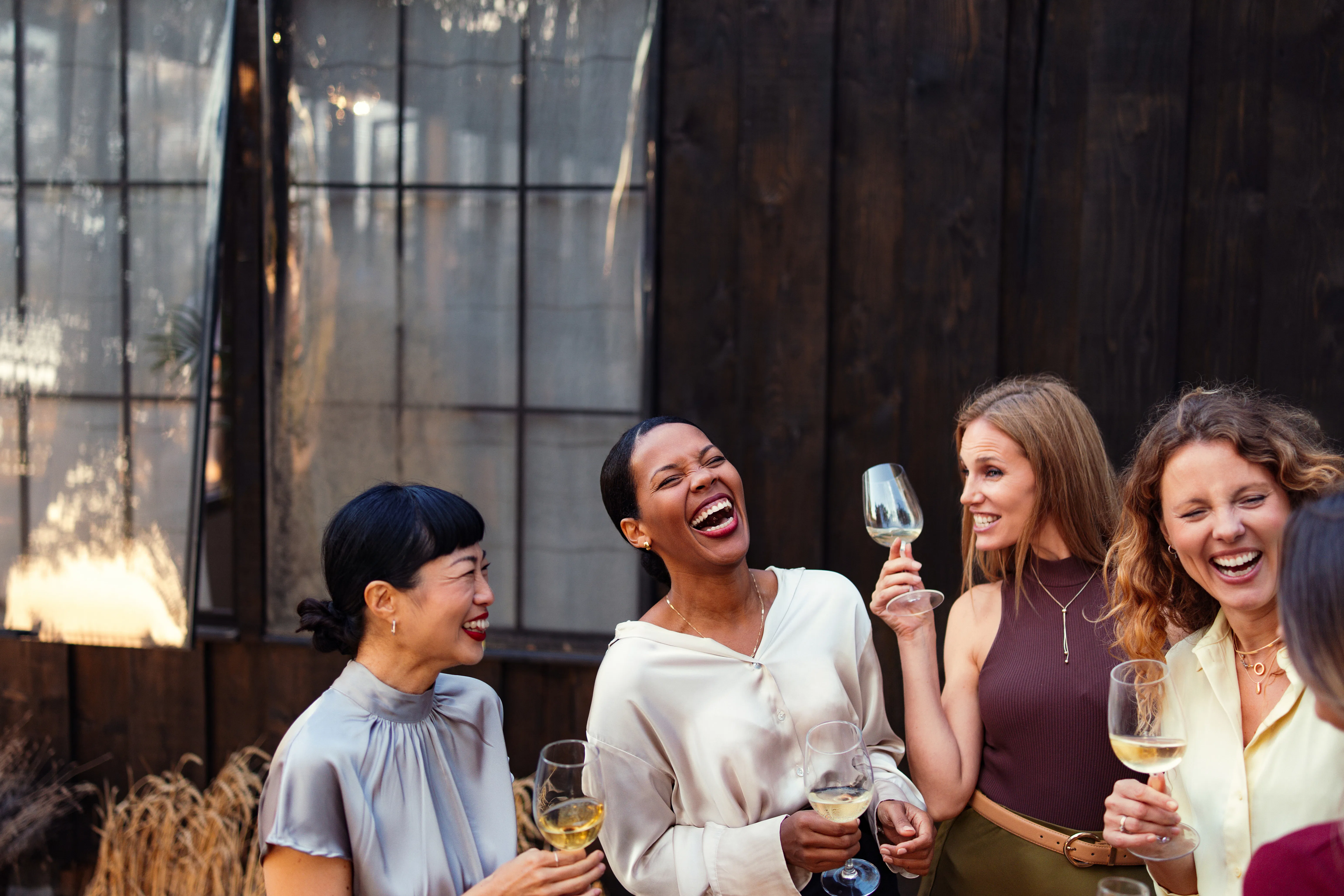 A diverse group of women laugh and socialize outdoors at a casual corporate event, holding wine glas...