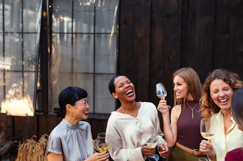 A diverse group of women laugh and socialize outdoors at a casual corporate event, holding wine glas...