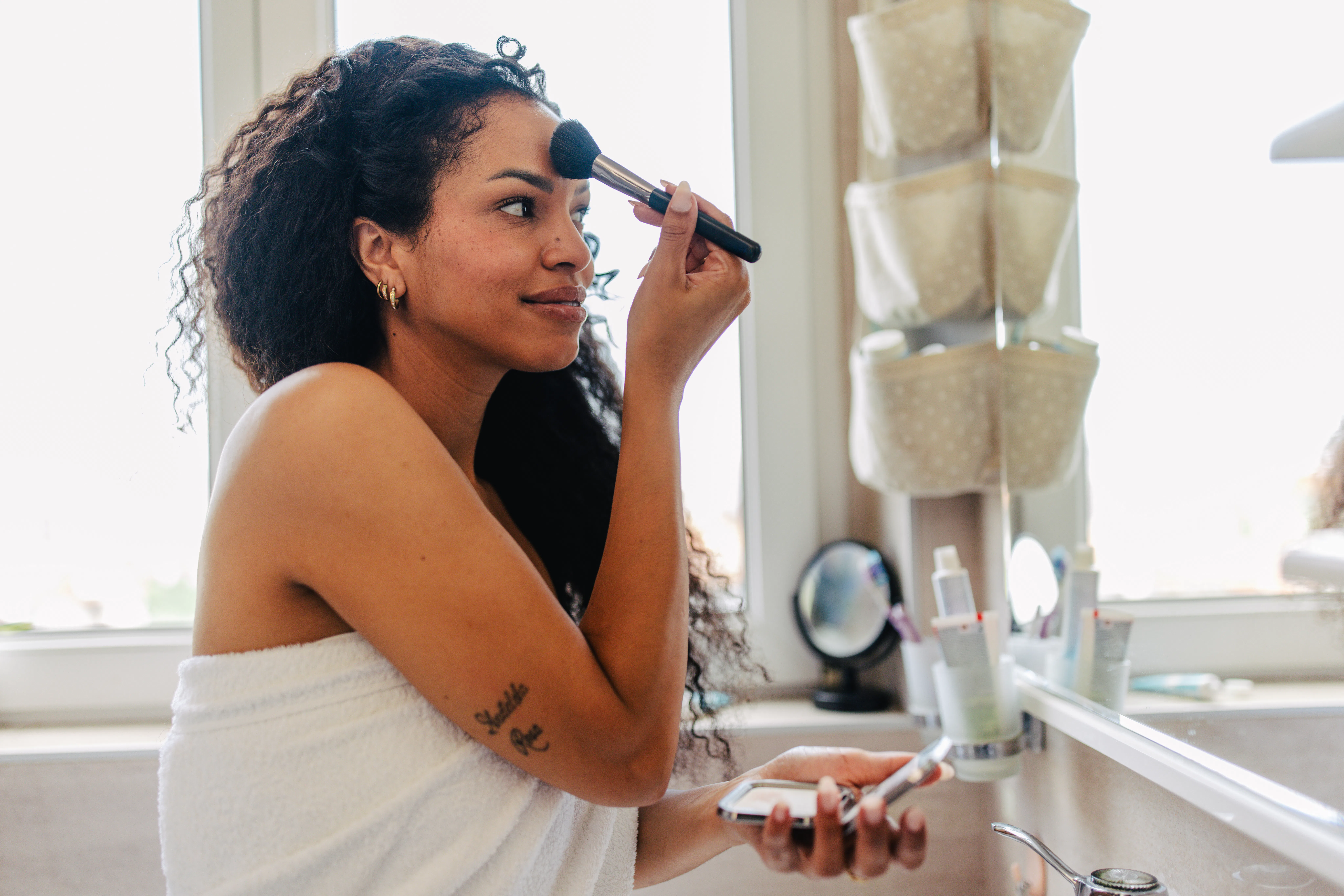 A woman with curly hair applies makeup in a bright, modern bathroom, surrounded by beauty products, ...