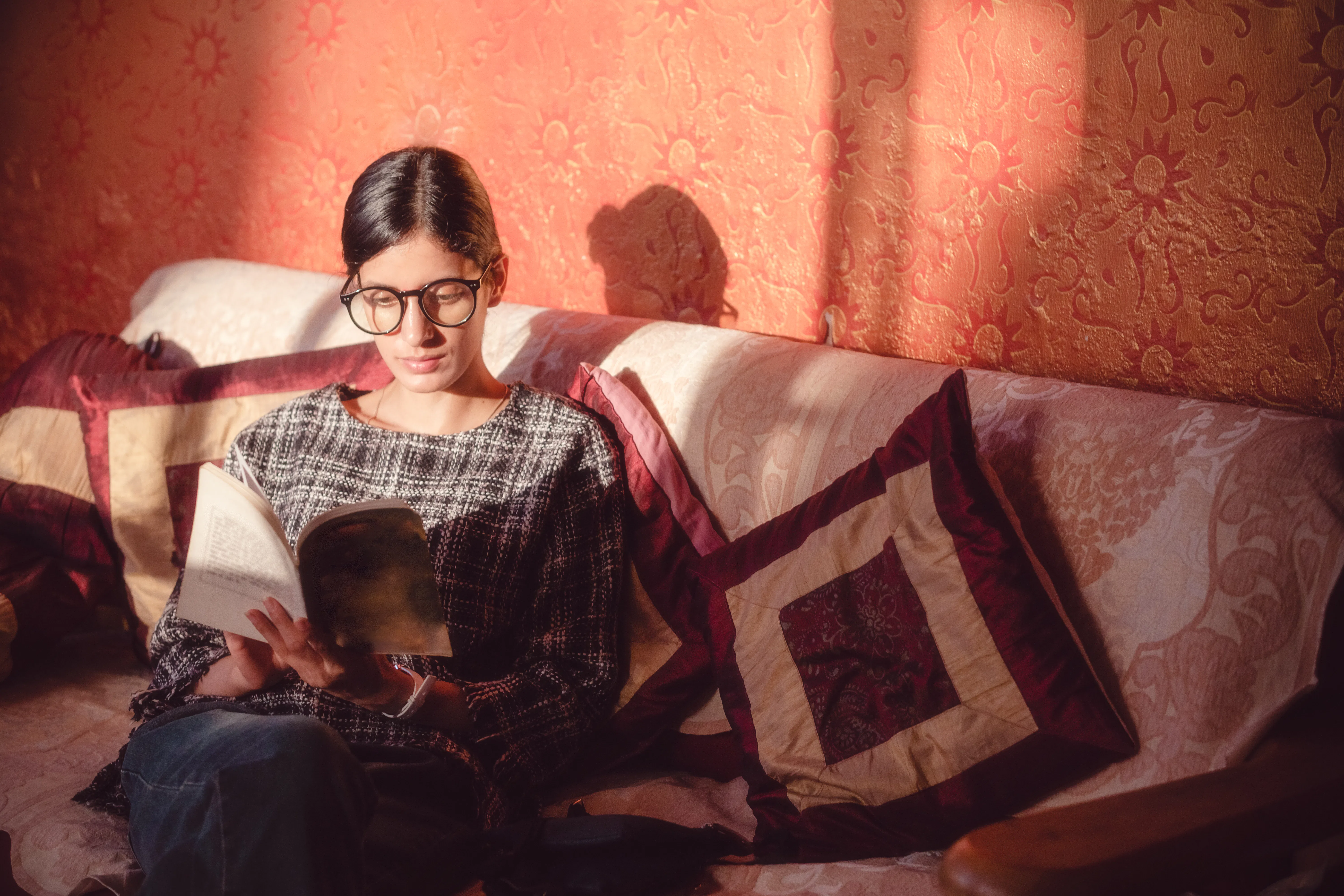 A focused young adult woman sits comfortably on a sofa, deeply engrossed in reading a book. She wear...