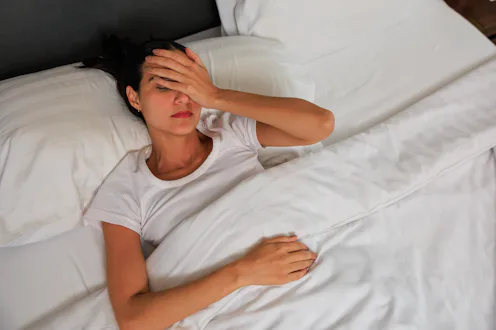 A tired and unwell young woman lies in bed with her eyes closed, holding her hand to her forehead in...