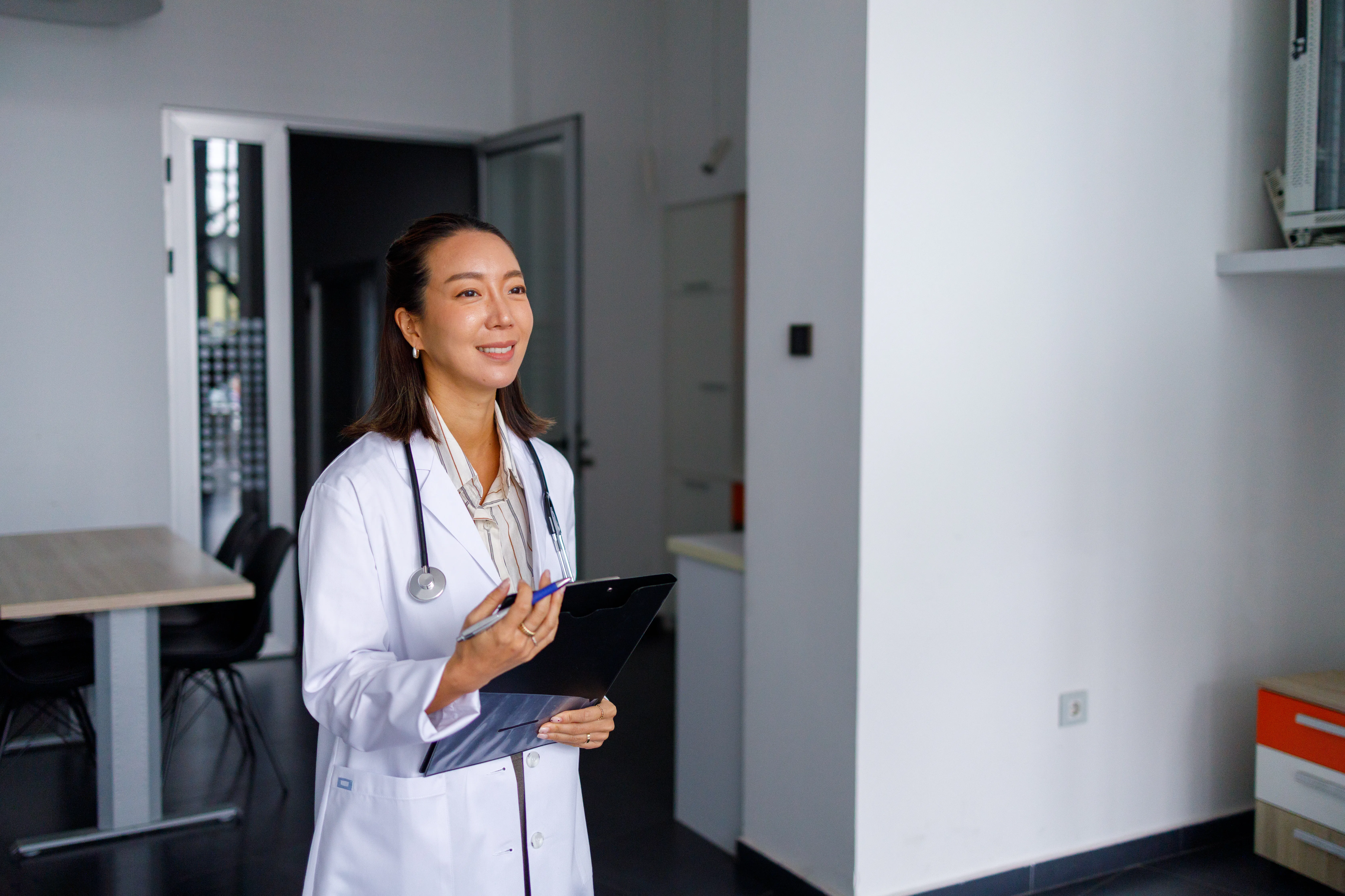 Female doctor in lab coat with stethoscope smiling while holding a clipboard and pen