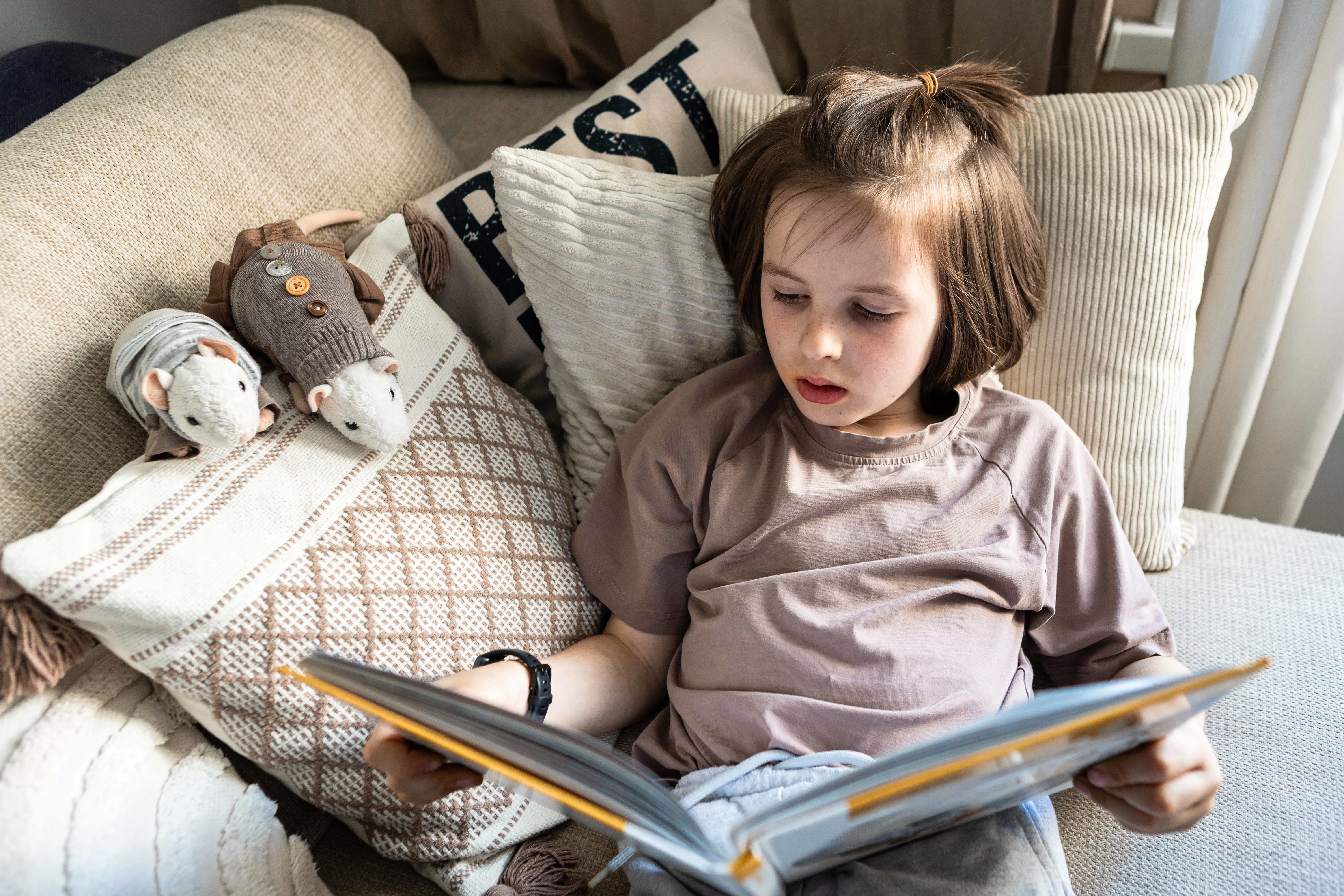 Young child sits among cushions reading a book at home in a quiet everyday living room scene.