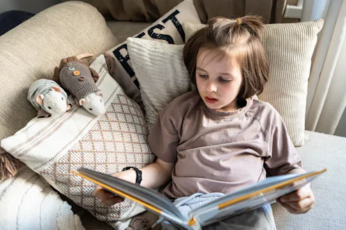 Young child sits among cushions reading a book at home in a quiet everyday living room scene.
