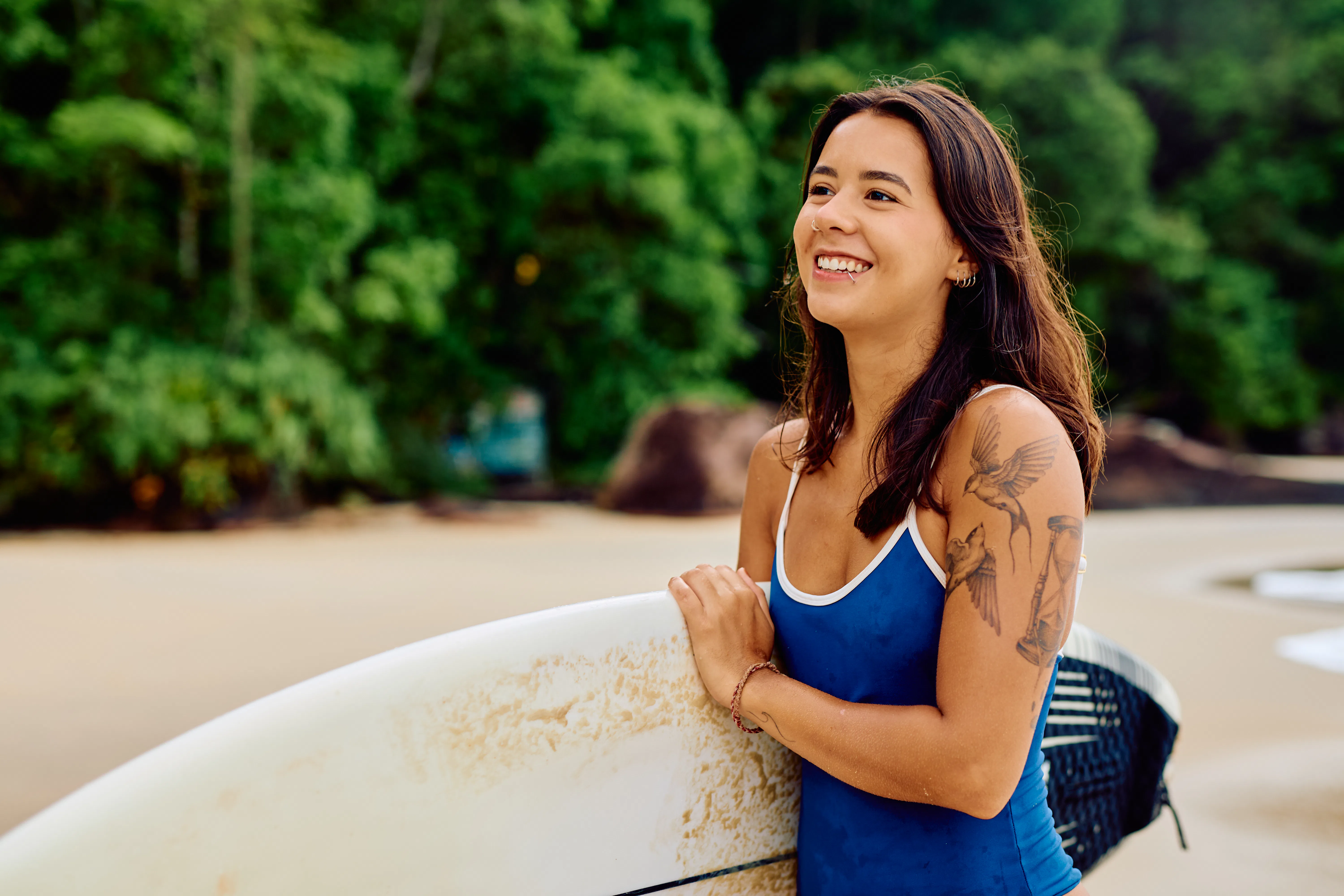 Young adult woman with a tattoo on her arm holding a surfboard by the ocean, enjoying a summer day o...