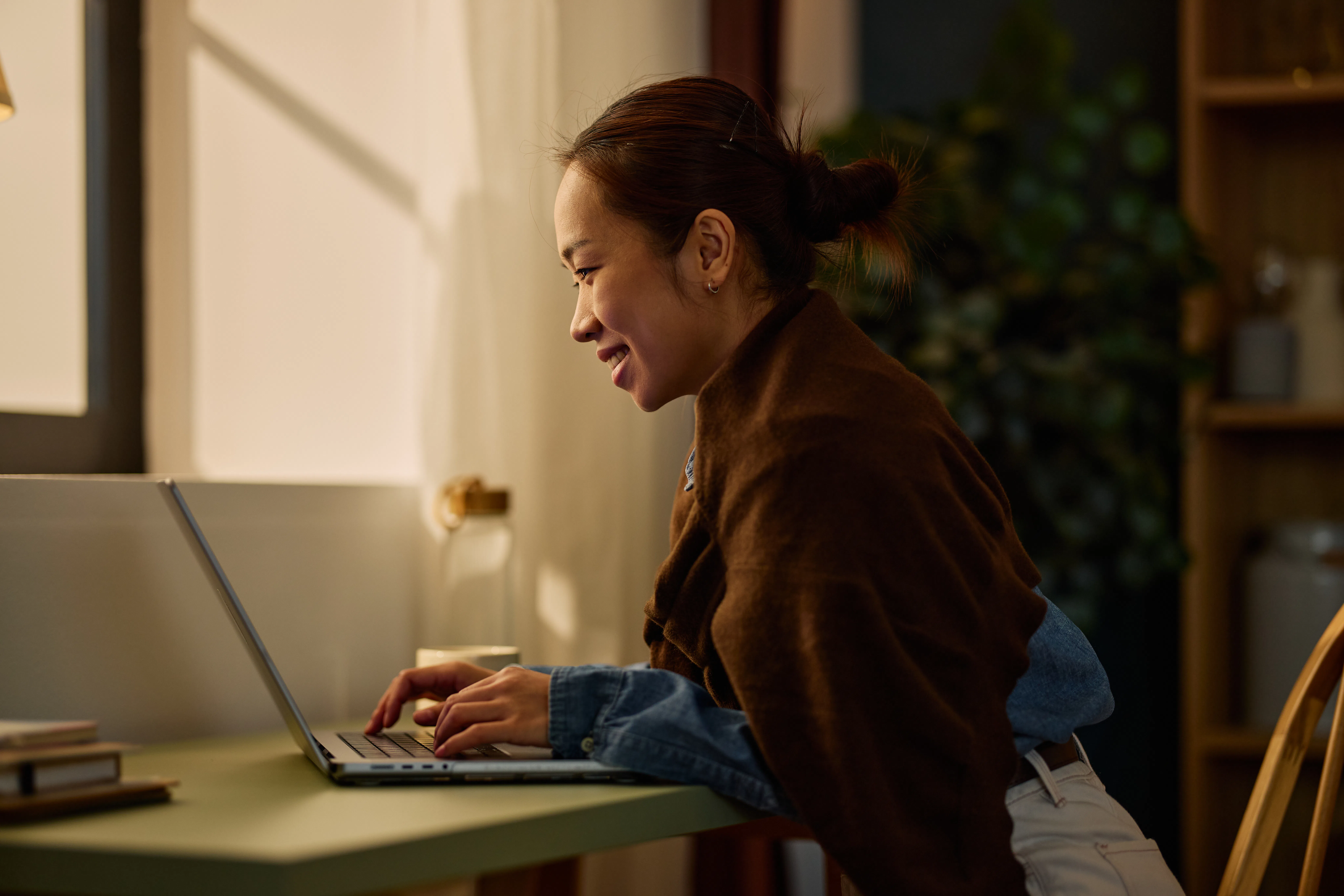Smiling Asian woman typing on laptop, working from home in a cozy evening setting. Remote work and t...