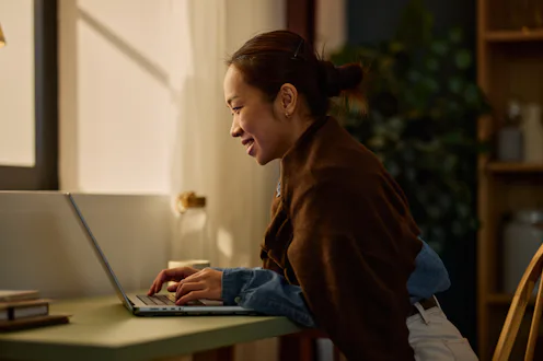 Smiling Asian woman typing on laptop, working from home in a cozy evening setting. Remote work and t...