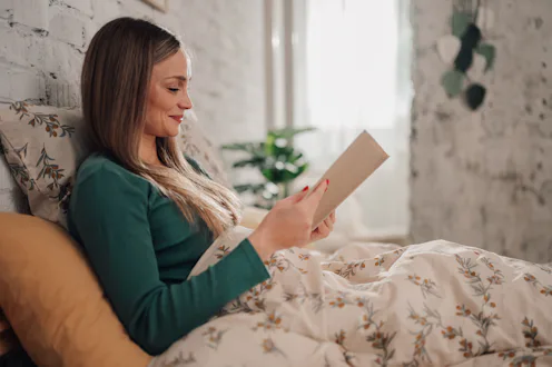 Young woman in her twenties lounging in bed in the morning, engrossed in a book. Embracing a serene ...