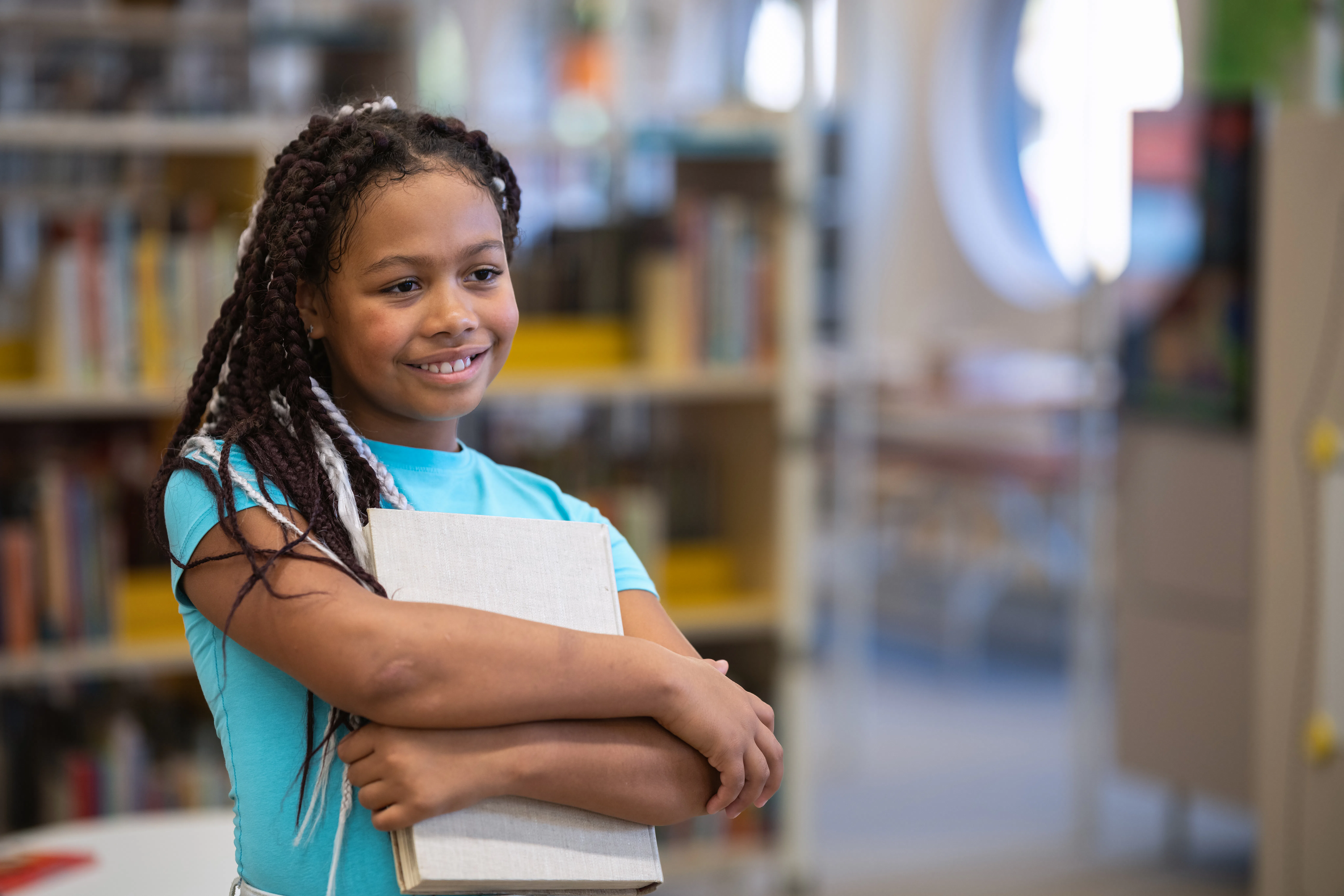 Dark-skinned mixed race girl in elementary school standing with a smile in library with book.