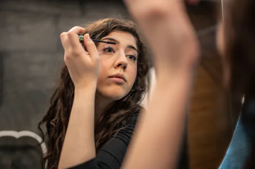 Young woman concentrates while applying mascara in front of a mirror during her evening makeup routi...