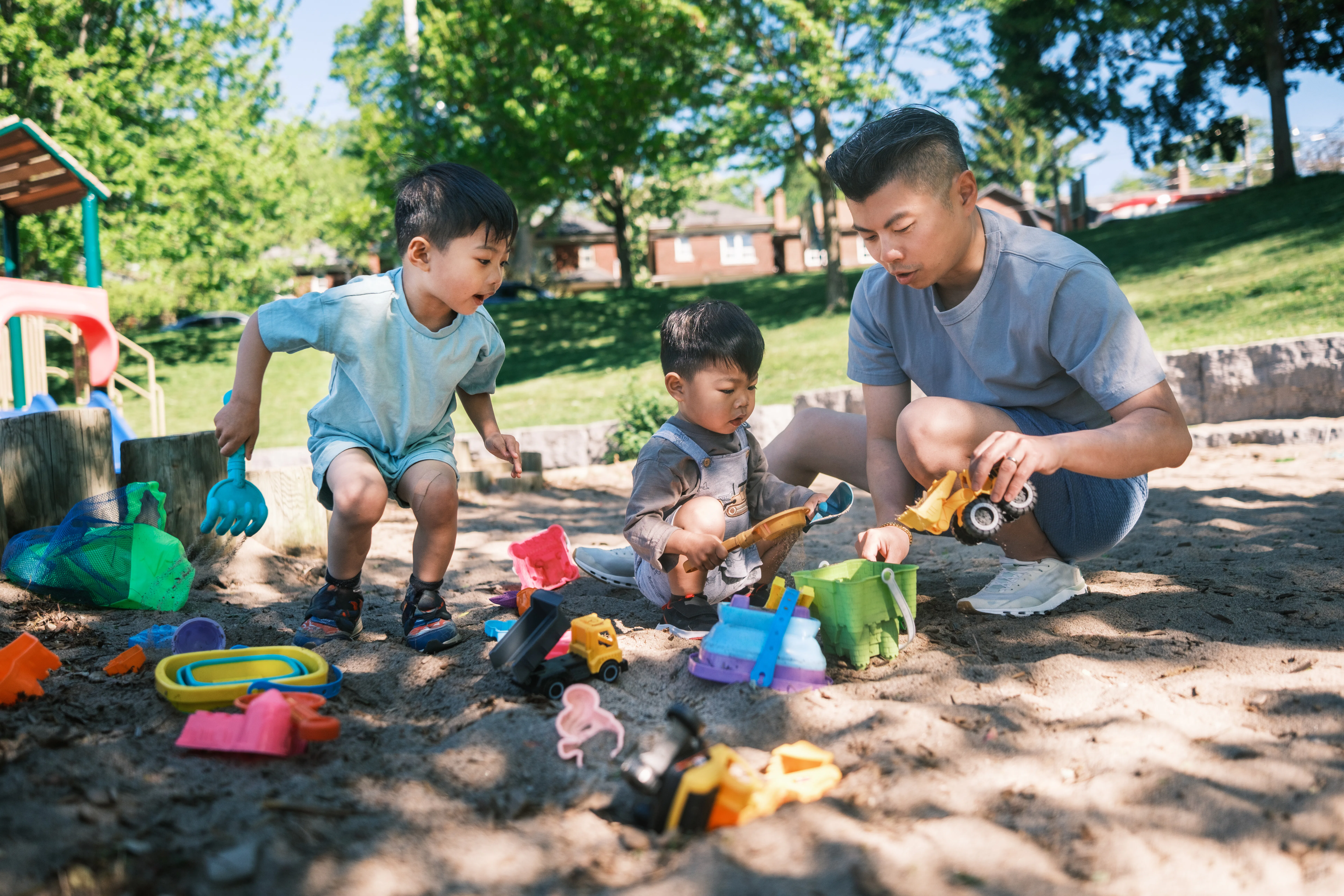 Young father and two sons playing in the sandbox  at the park. All dressed in casual summer clothing...