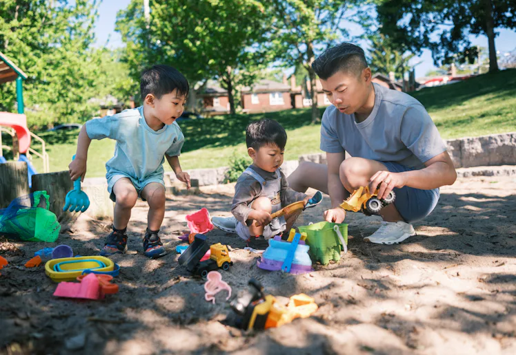Young father and two sons playing in the sandbox at the park. All dressed in casual summer clothing...