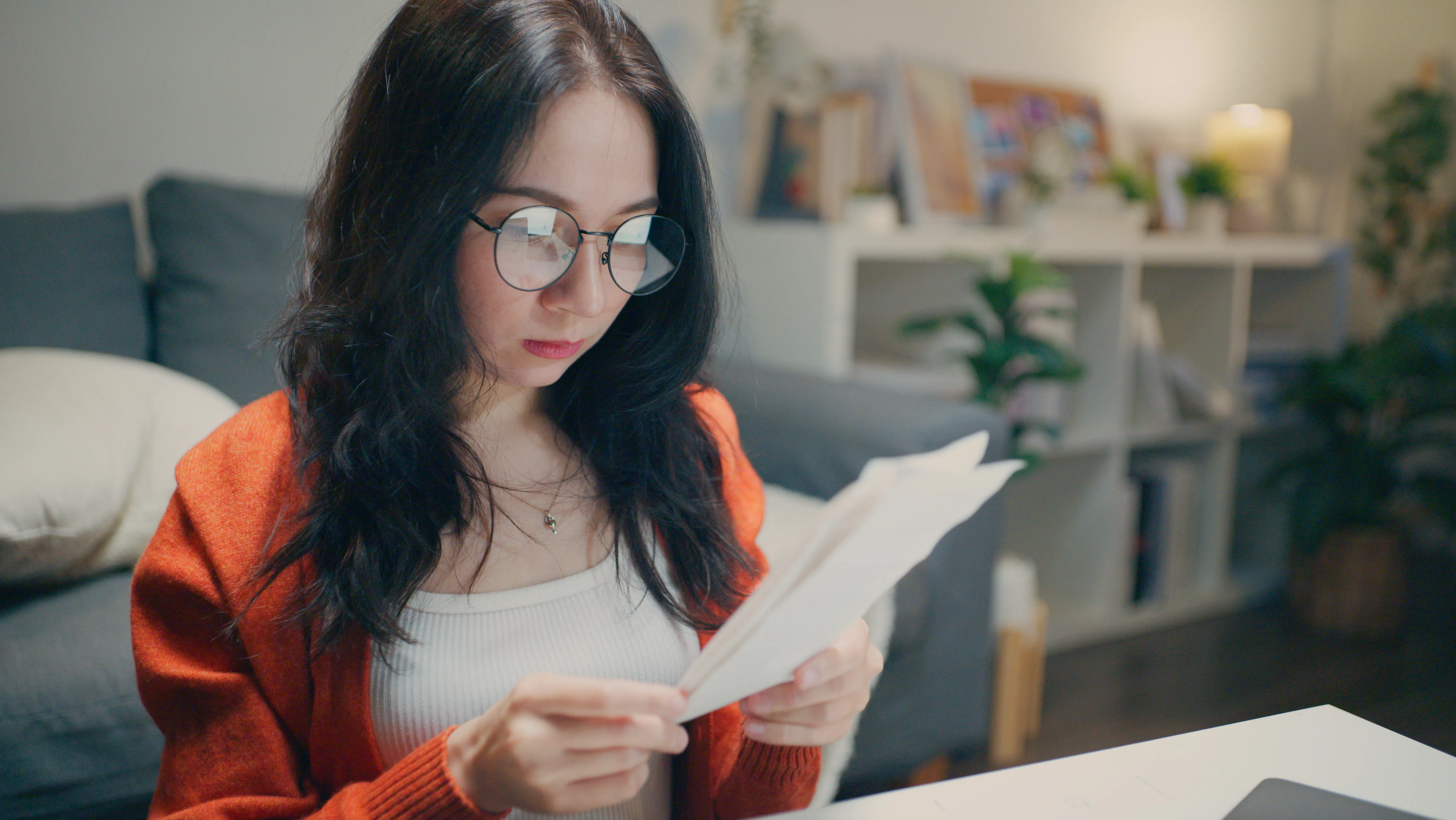 Serious Asian woman wearing glasses, holding and examining bills or financial document at desk at ni...