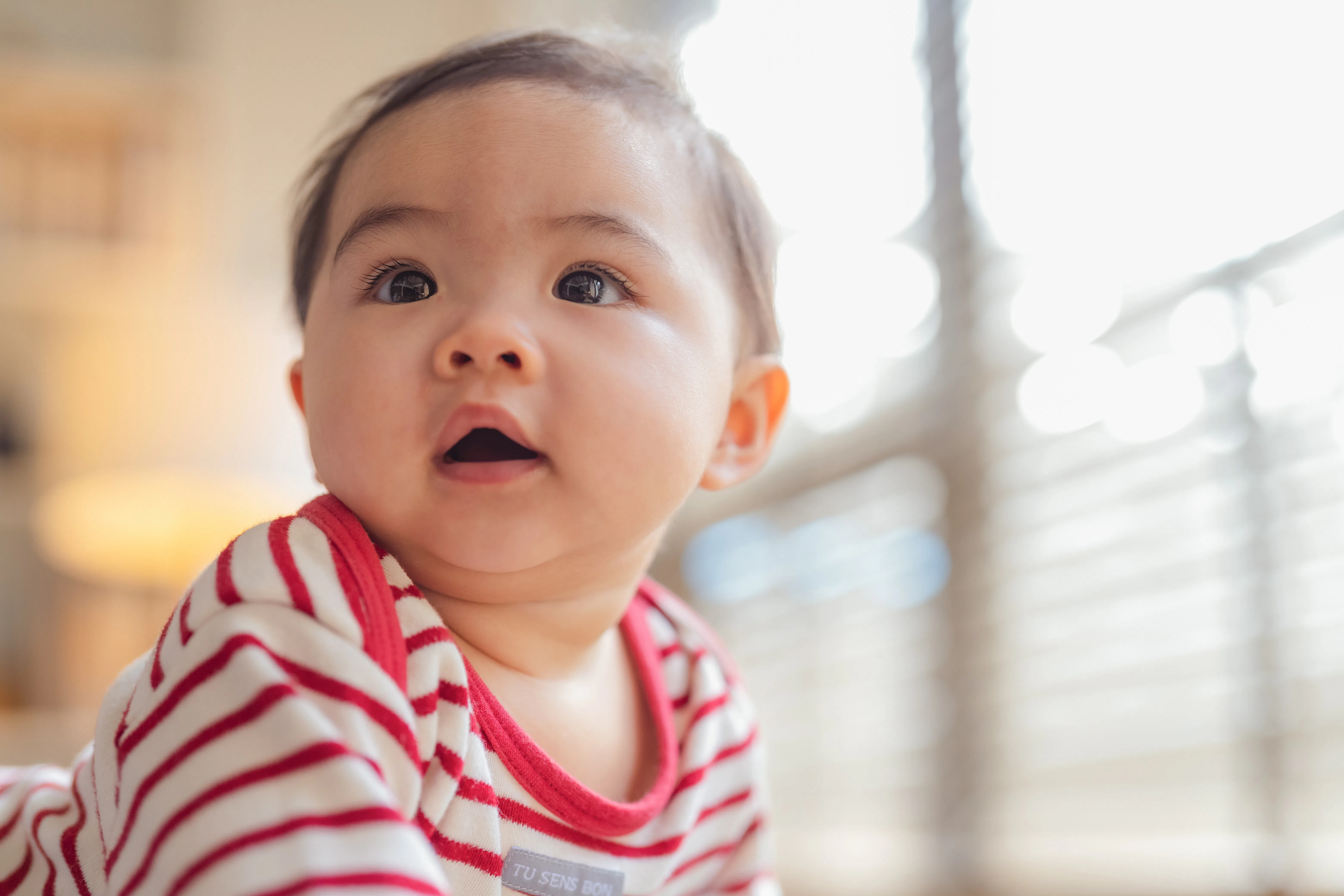 Happy Asian baby girl practices standing and balancing on a soft stool ottoman at home, developing b...