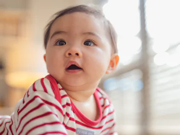 Happy Asian baby girl practices standing and balancing on a soft stool ottoman at home, developing b...