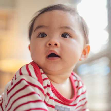 Happy Asian baby girl practices standing and balancing on a soft stool ottoman at home, developing b...