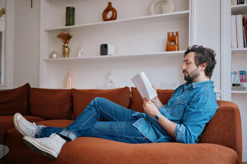 Young man reading book while relaxing on sofa at home in Paris