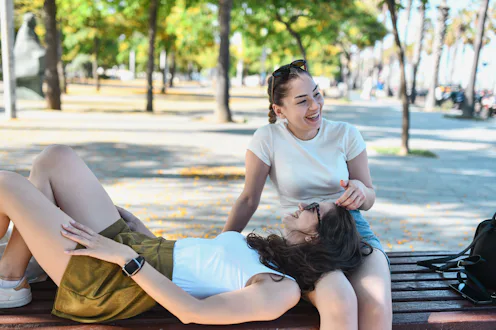 Two young women relax and laugh together on a park bench in Barcelona, enjoying a sunny, peaceful af...