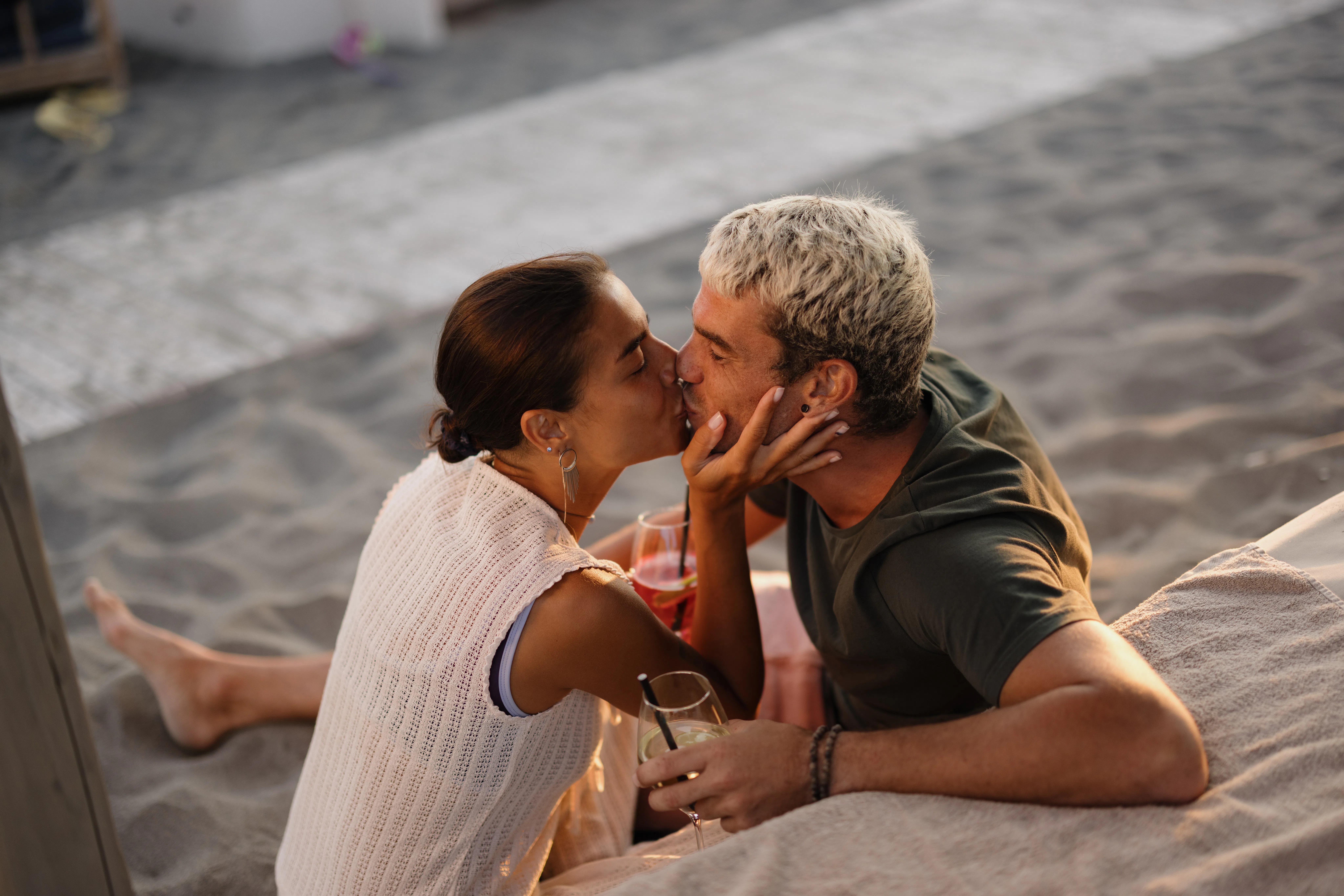 Young heterosexual couple sharing an intimate kiss during sunset on a sandy beach while enjoying coc...