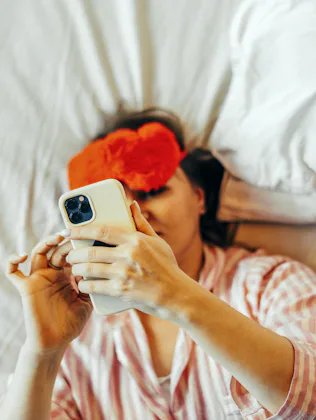 A woman in the bed with red Eye Mask and mobile phone in hand.