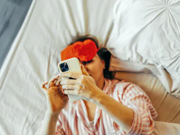 A woman in the bed with red Eye Mask and mobile phone in hand.