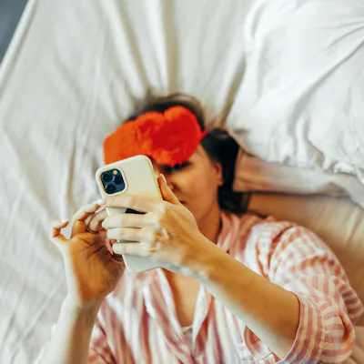 A woman in the bed with red Eye Mask and mobile phone in hand.