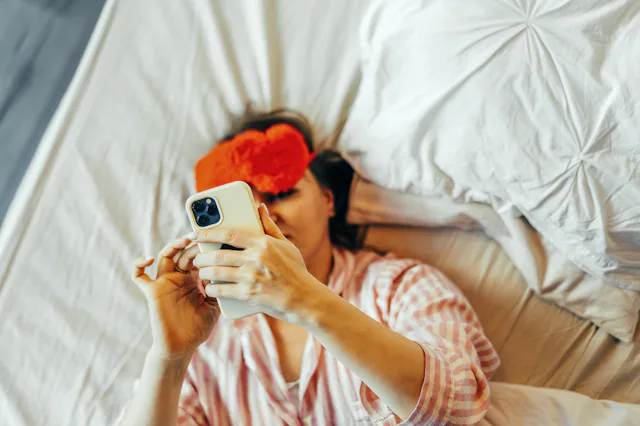 A woman in the bed with red Eye Mask and mobile phone in hand.