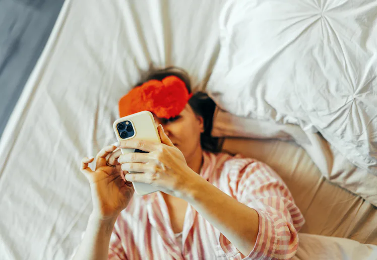 A woman in the bed with red Eye Mask and mobile phone in hand.
