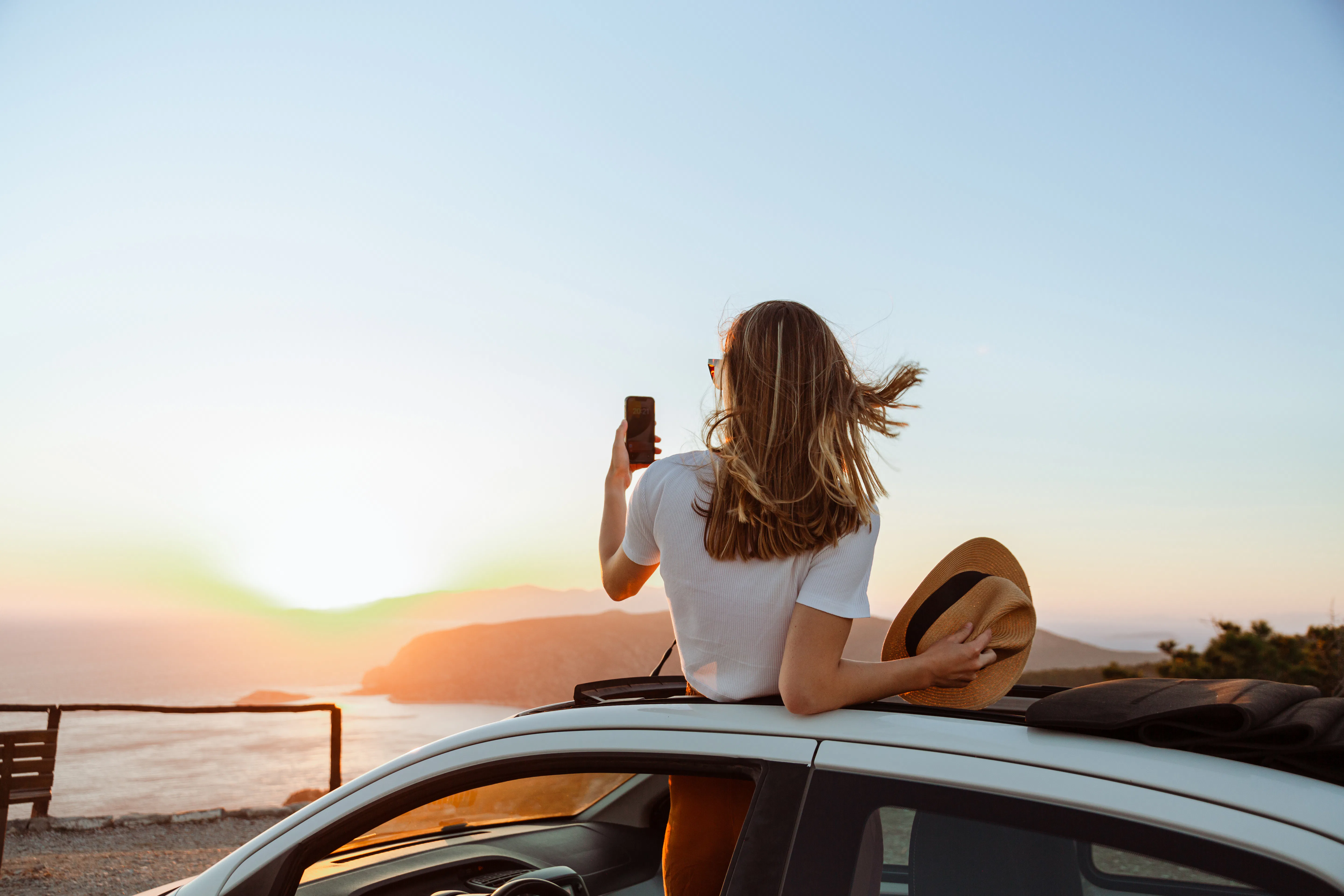 A photo of an unrecognizable woman looking at the sea and mountains from the opened roof of her car....