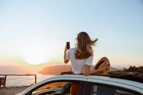 A photo of an unrecognizable woman looking at the sea and mountains from the opened roof of her car....