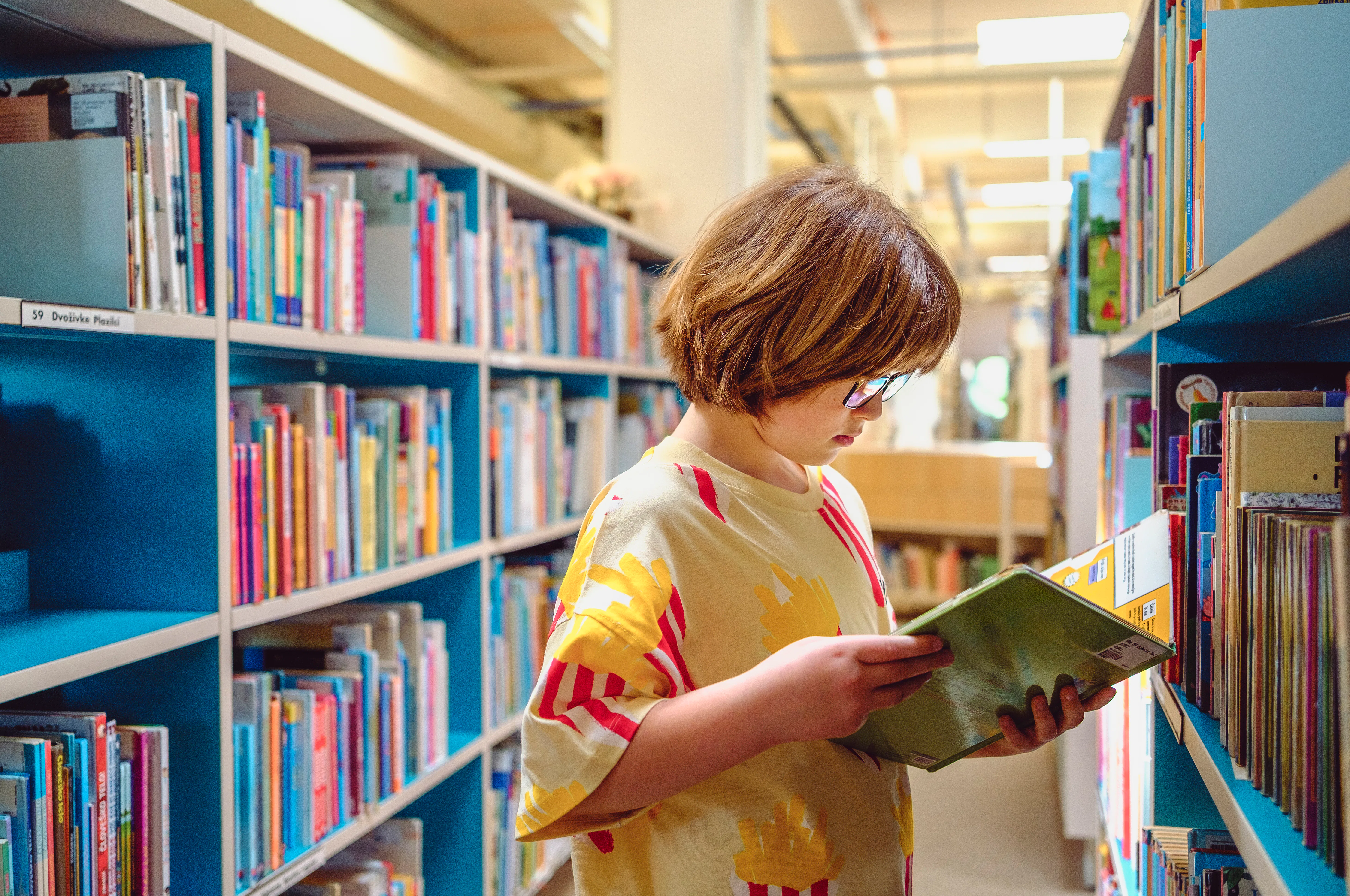 A Caucasian boy wearing a yellow and red striped shirt stands between tall library shelves, absorbed...