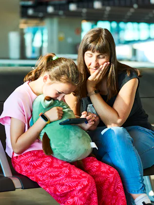 Mother and daughter embracing while playing a video game on a smartphone, relaxing during a long wai...