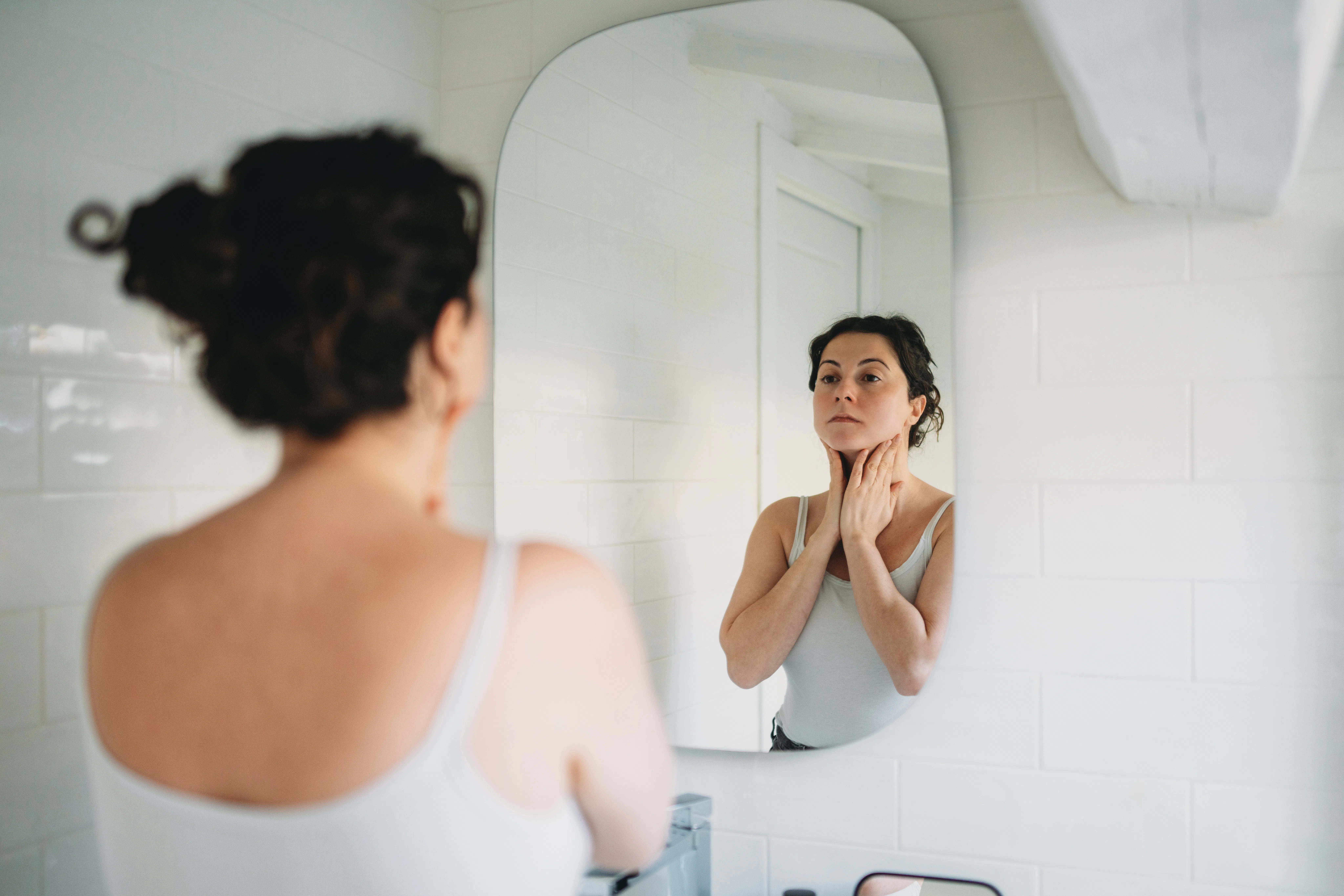 Woman in a bathroom, gazing into the mirror while gently touching her neck, reflecting on her skinca...