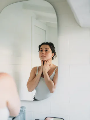 Woman in a bathroom, gazing into the mirror while gently touching her neck, reflecting on her skinca...