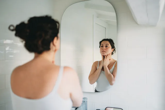 Woman in a bathroom, gazing into the mirror while gently touching her neck, reflecting on her skinca...