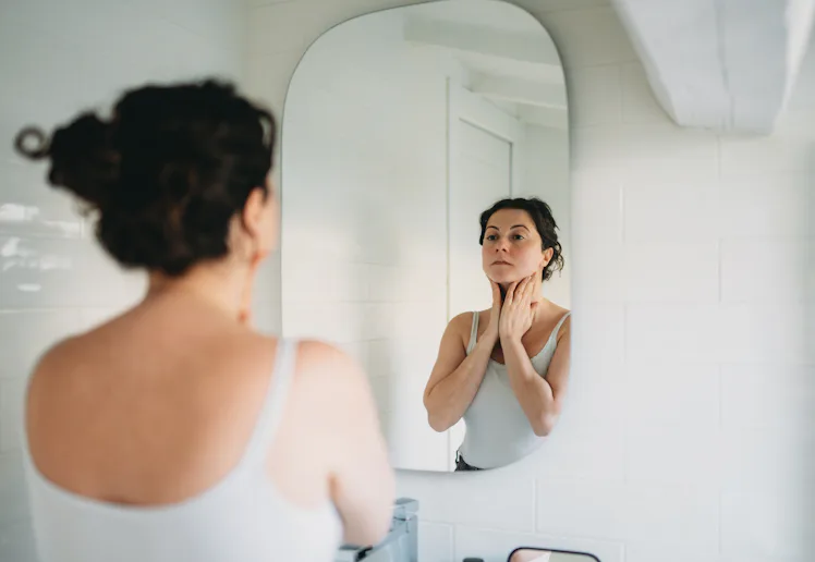 Woman in a bathroom, gazing into the mirror while gently touching her neck, reflecting on her skinca...