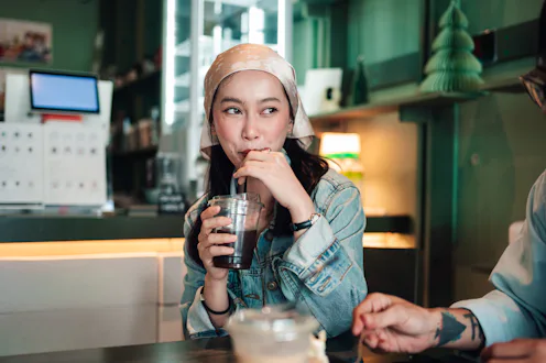 Young asian woman drinking iced coffee with a straw, observing and chatting in a stylish, cozy coffe...