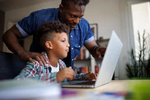 African American father helping his young son during an online learning session at home. Caring dad ...