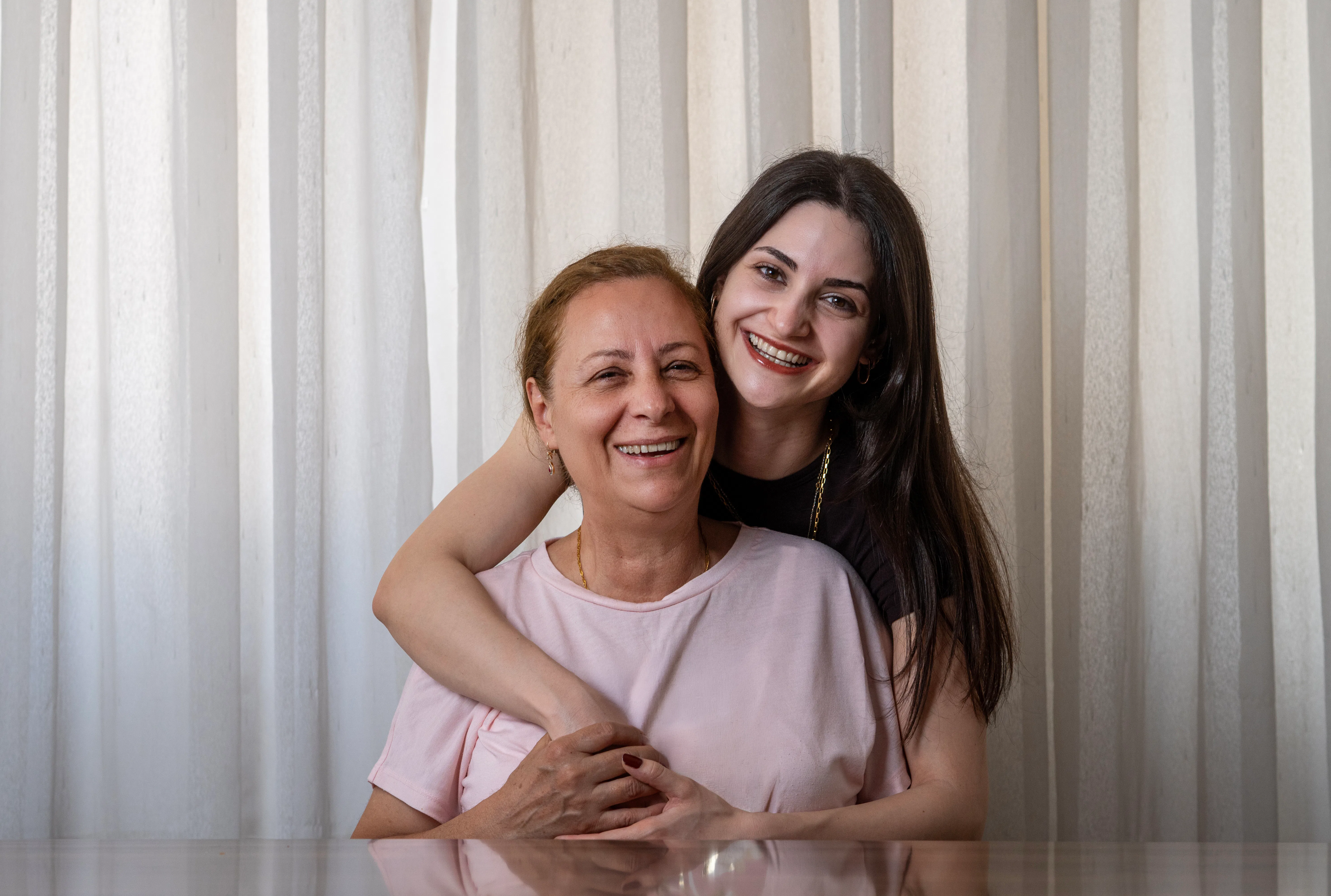 Young woman embracing her mother from behind with a big smile against curtains
