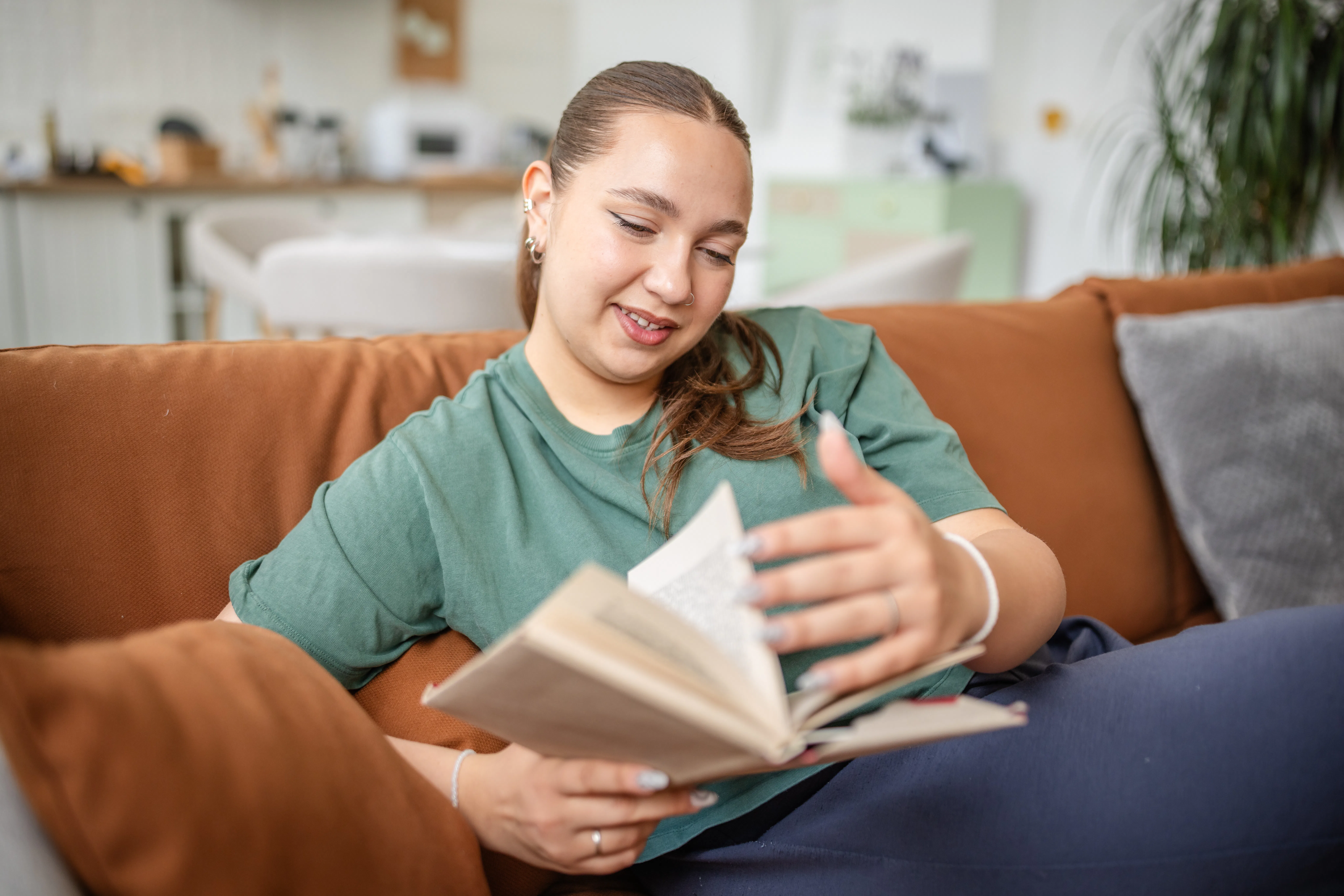 A plus-sized young woman sits comfortably on a couch, engrossed in a book. The bright living room fe...