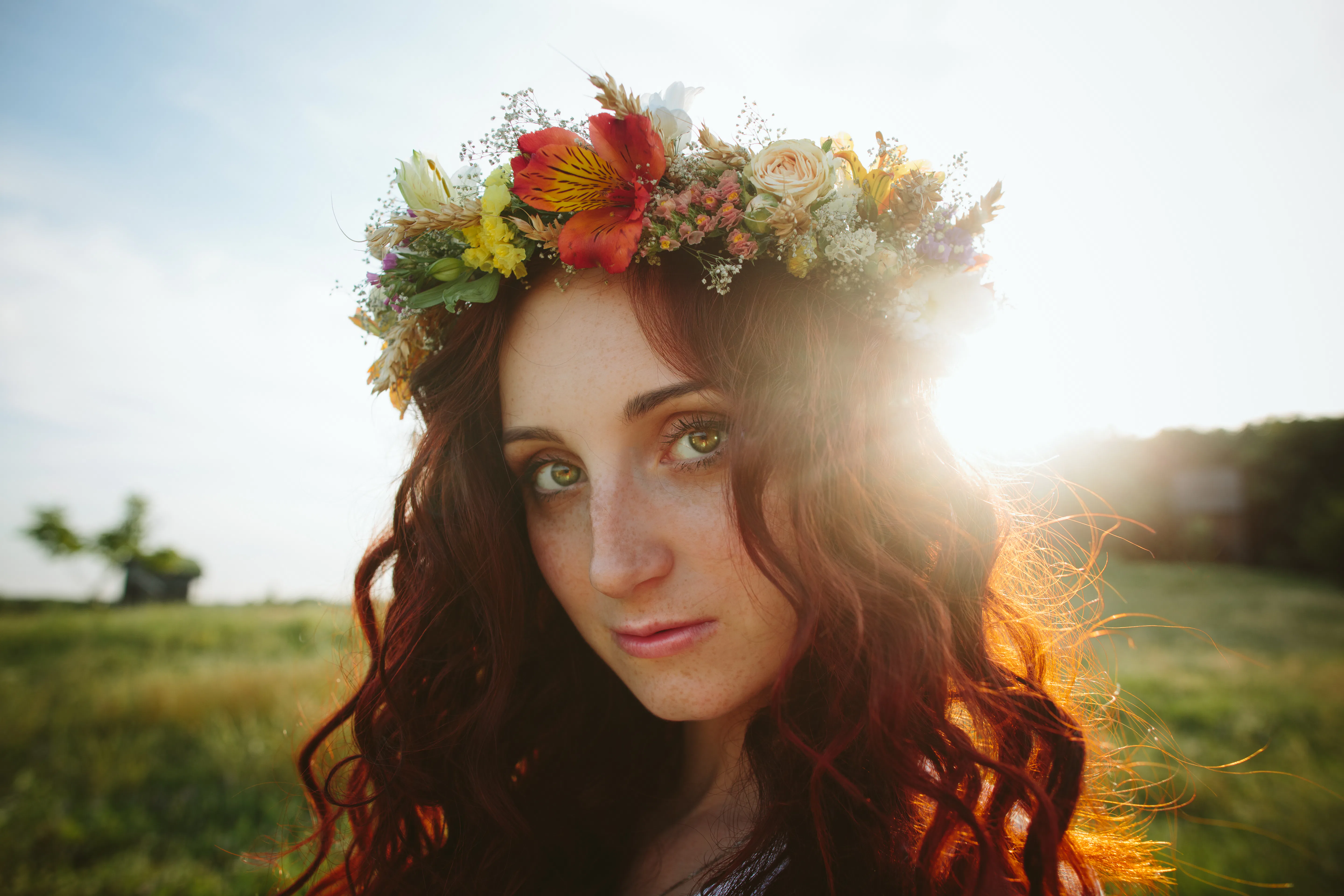 Young redhead woman with a spring wreath sits on the field grass against the background of old mills...