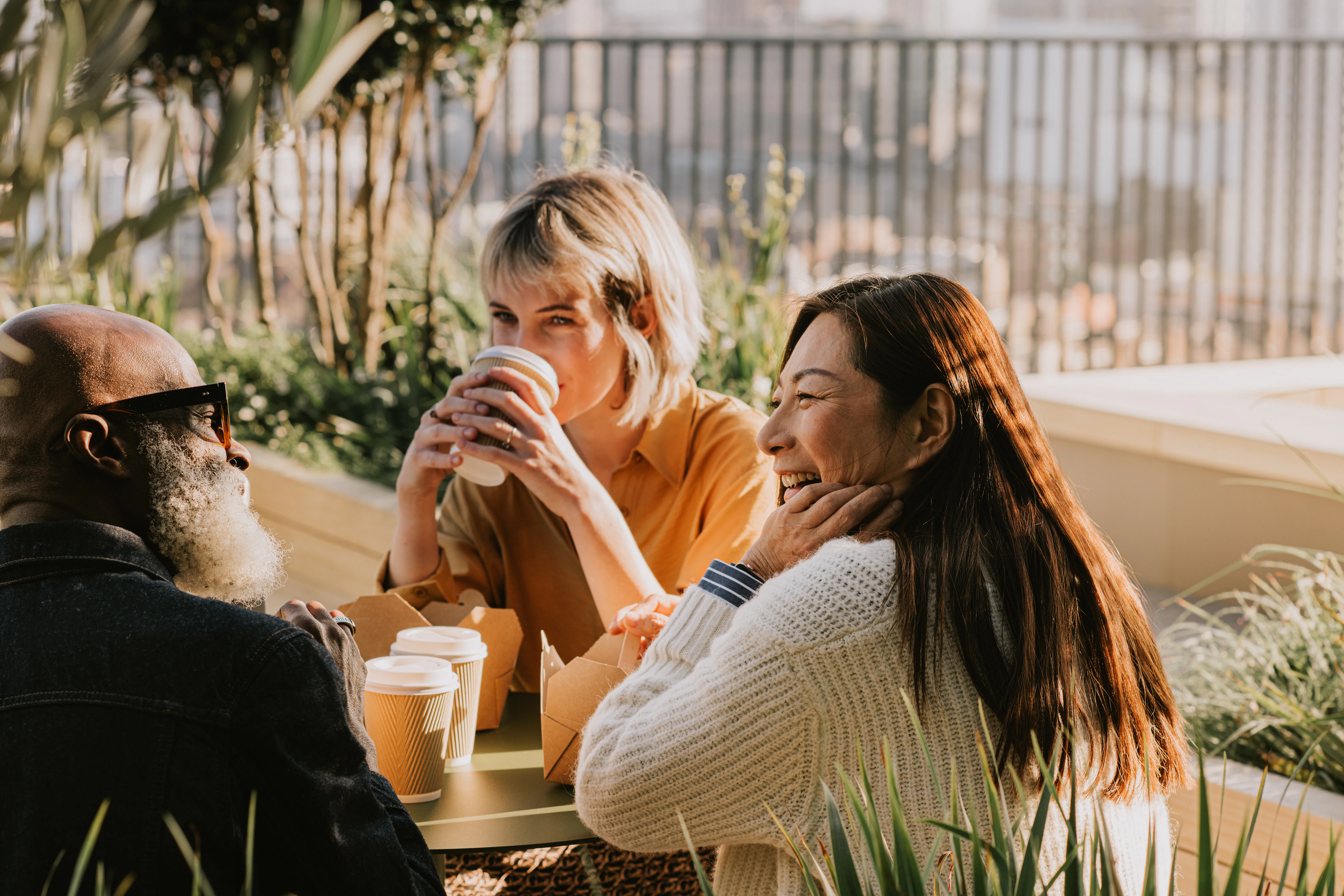 Three colleagues sit outside on a sunny day and enjoy a takeaway lunch and some hot drinks. They are...